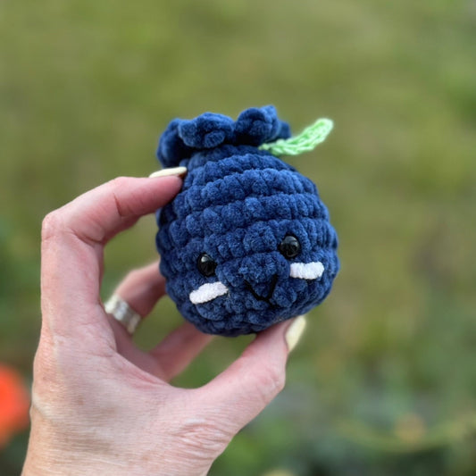 Hand holding a blue crochet fruit against a blurred natural background