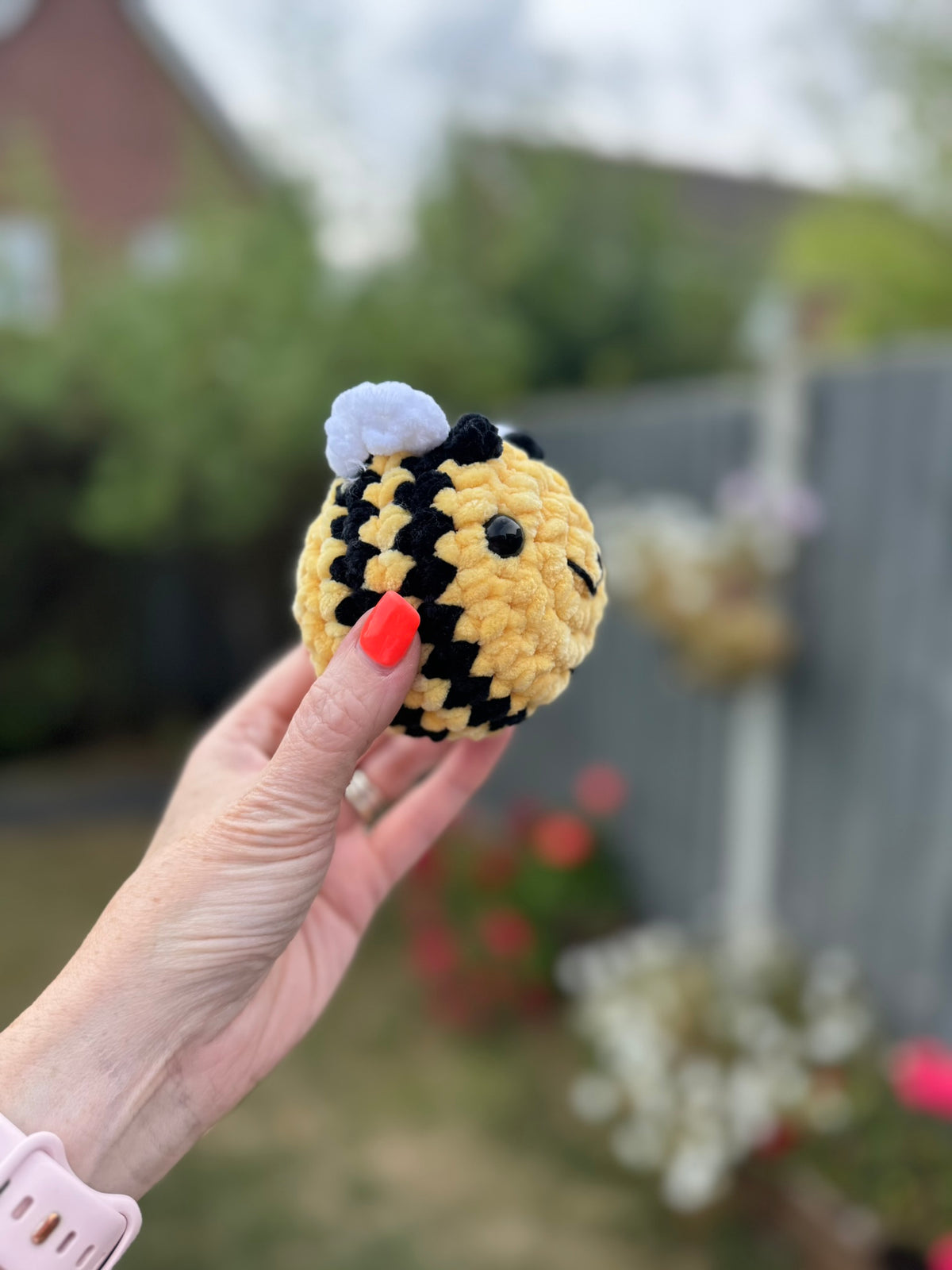 A hand with red nail polish holds a small crocheted bumblebee toy outdoors. The toy is yellow and black striped with a white fuzzy body and small black eyes.