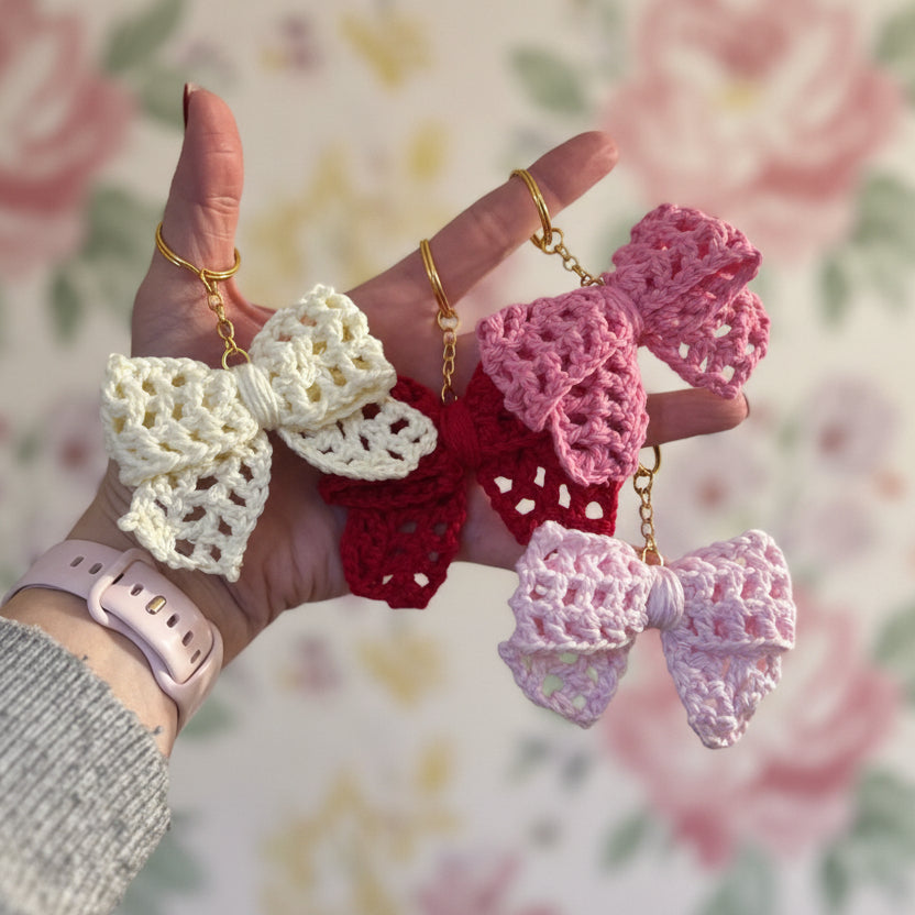 Hand holding three crocheted bow keychains in white, red, and pink against a floral background.