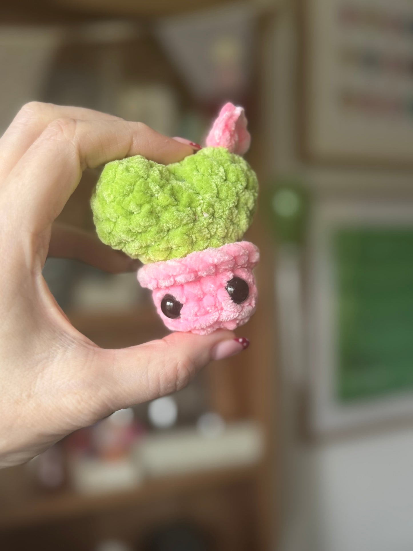 Hand holding a small crocheted green and pink creature toy against a blurred background