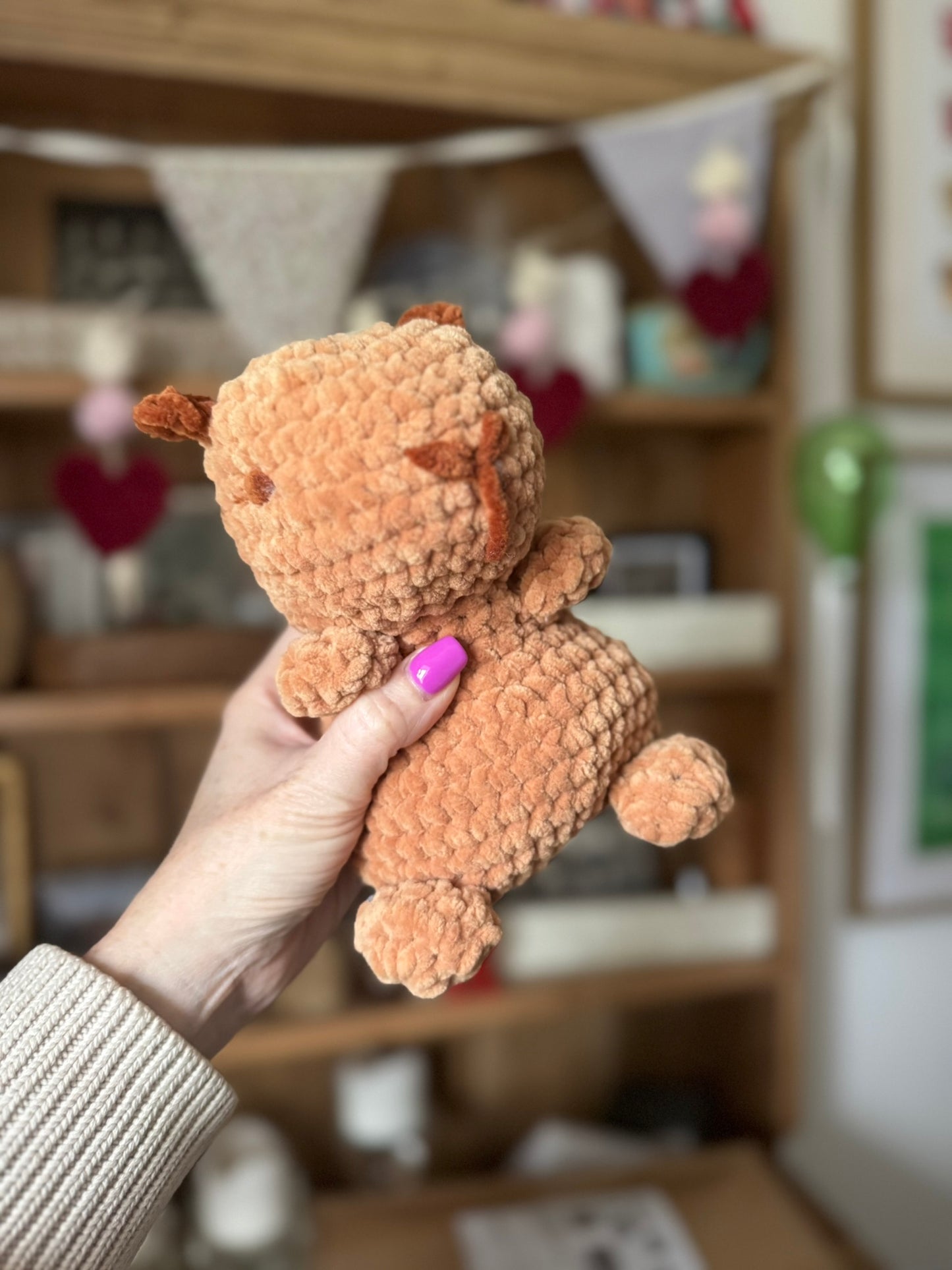 Hand holding a small brown teddy bear in front of a blurred indoor background