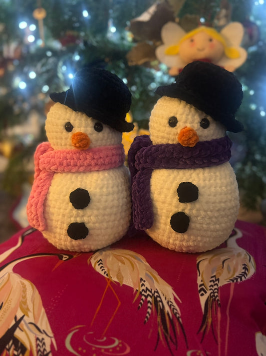 Two crochet snowmen with scarves in front of a decorated Christmas tree.
