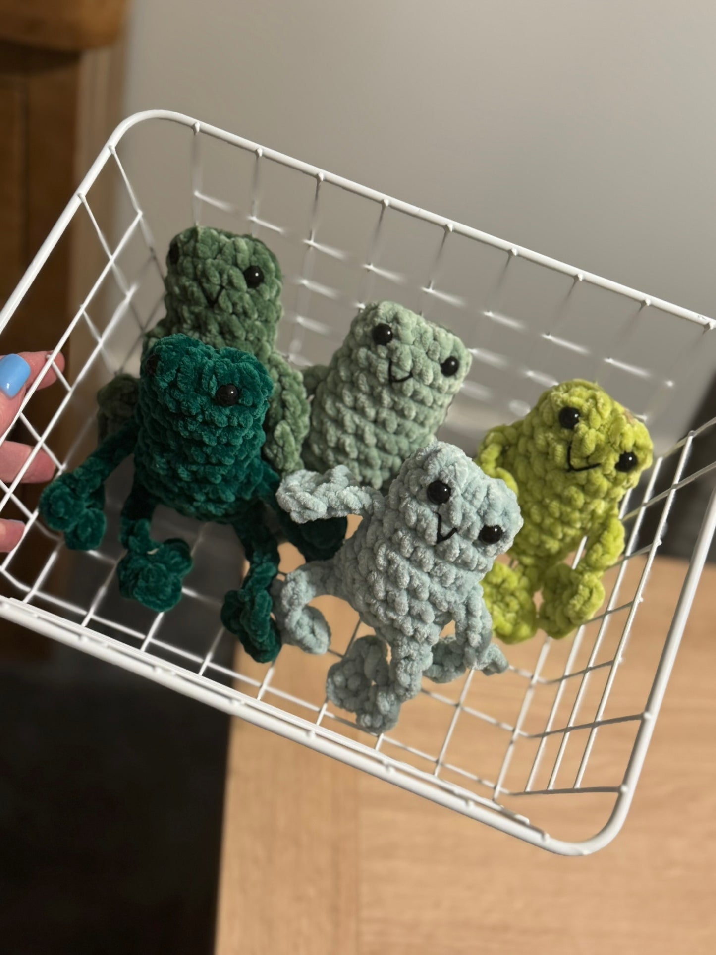 Five crocheted frog toys in a white metal basket on a wooden surface.
