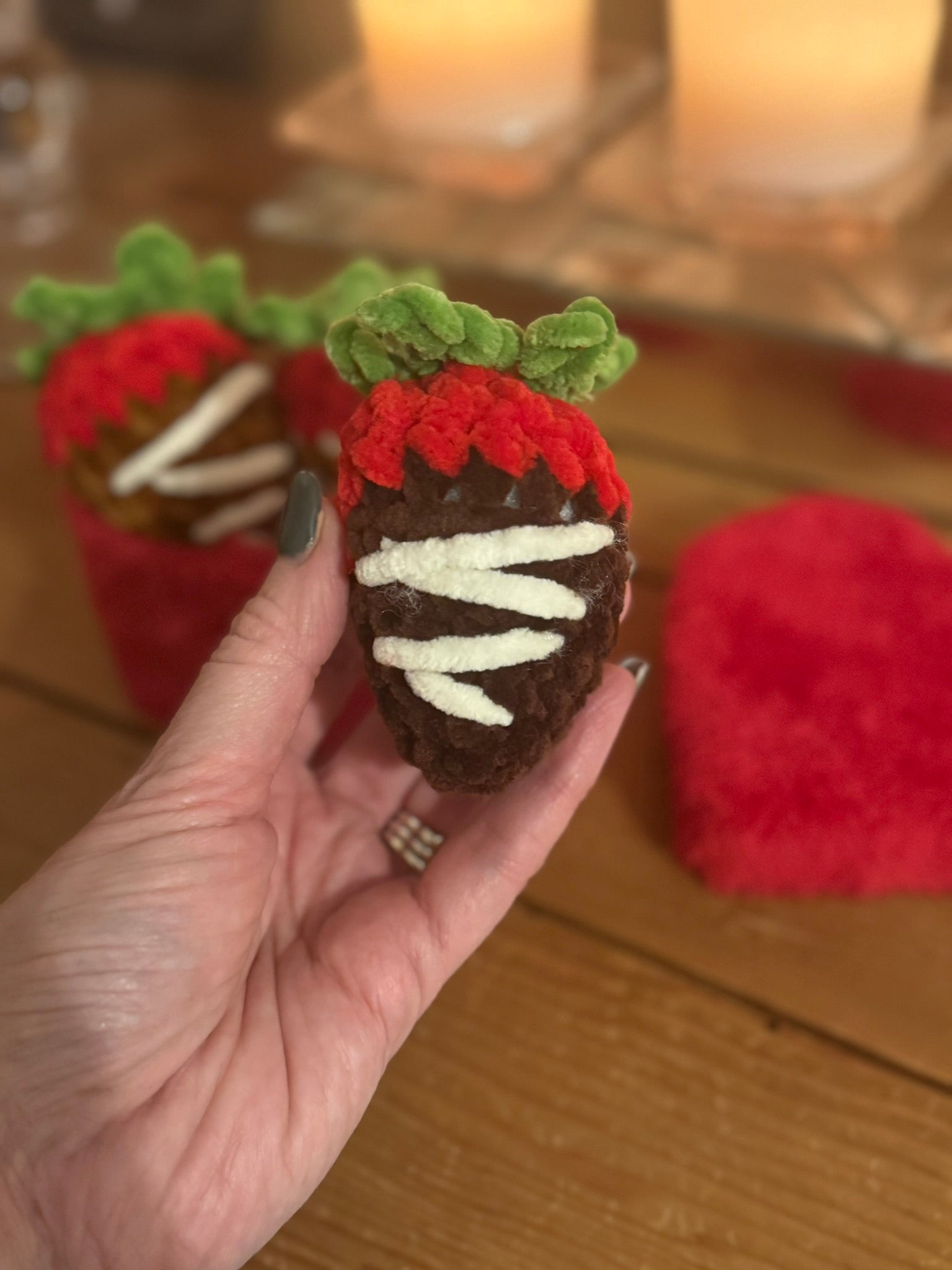 Hand holding a chocolate-covered strawberry-shaped treat with red and green decorations on a wooden surface.