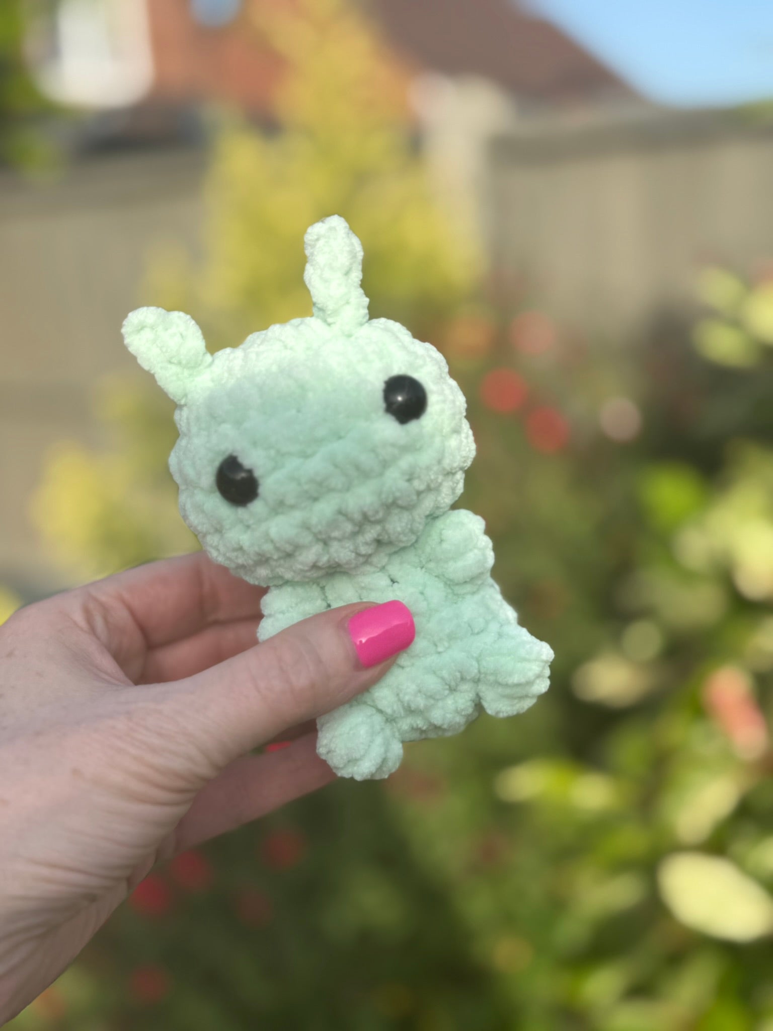 Small white plush toy held by a hand with pink nail polish against a blurred outdoor background.
