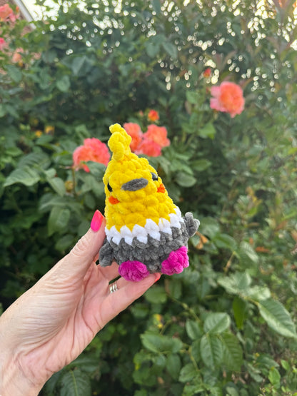A hand holds a small crocheted plush toy shaped like a yellow bird or chick with gray wings and pink feet, set against a blurred garden background with green leaves and pink flowers.