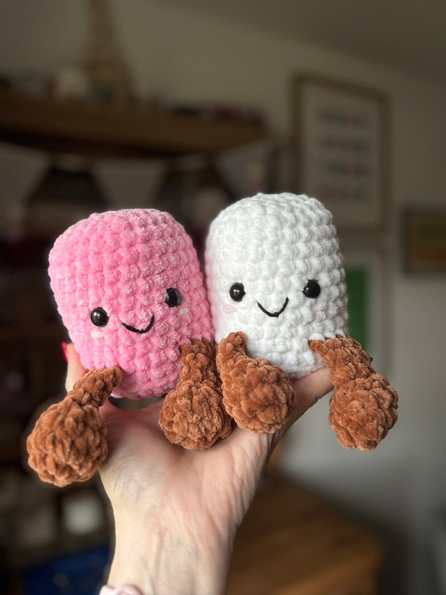 Two crocheted marshmallow toys, one pink and one white, held in a hand with a blurred background.