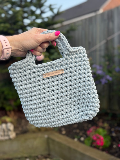 Hand holding a light blue knitted bag with a brand label against a blurred outdoor background