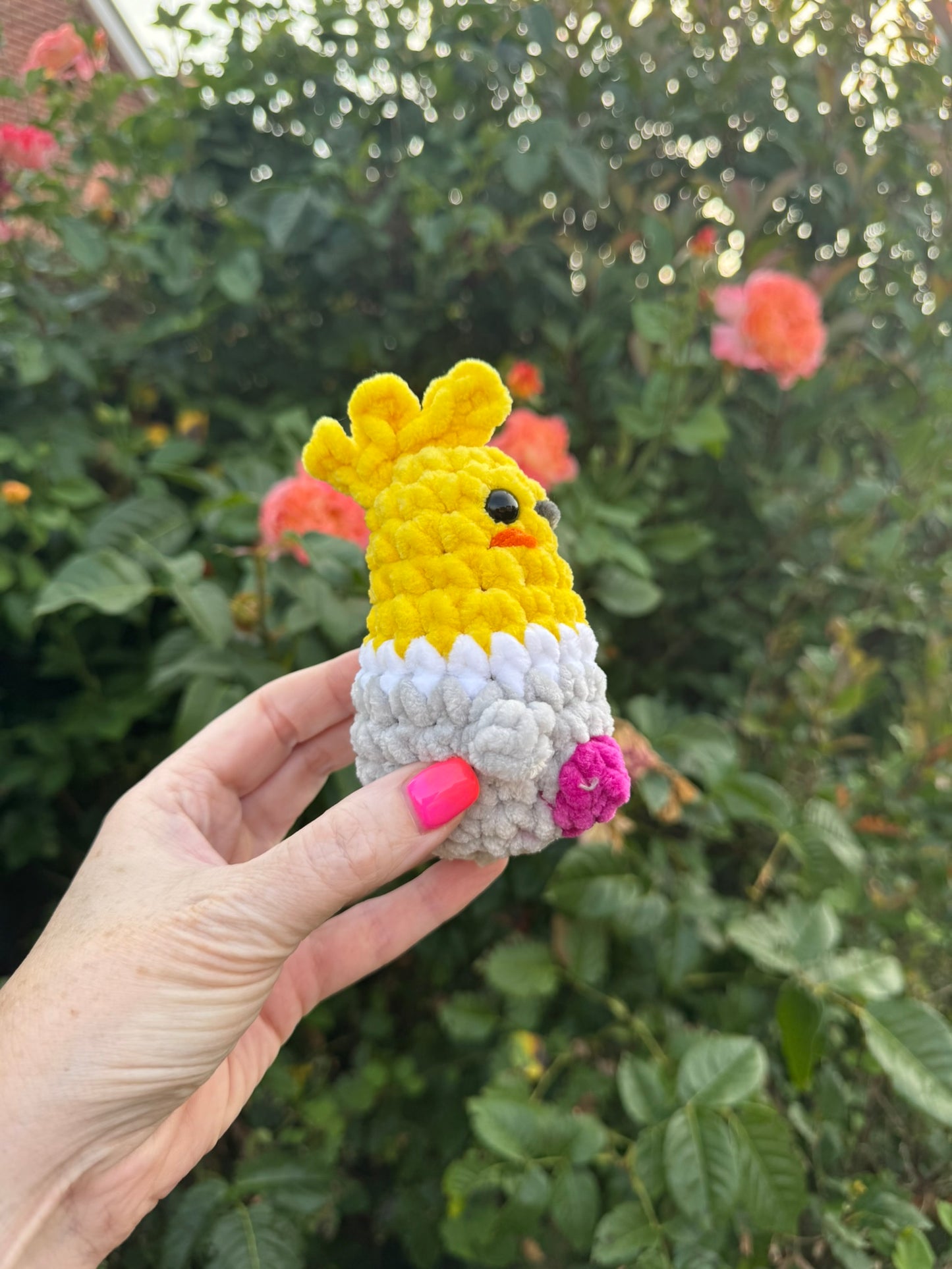 A hand with pink nail polish holds a small crocheted chick toy in front of a blurred garden background with green leaves and pink flowers. The toy is yellow on top, white on the bottom, and has a small pink foot and black eye.