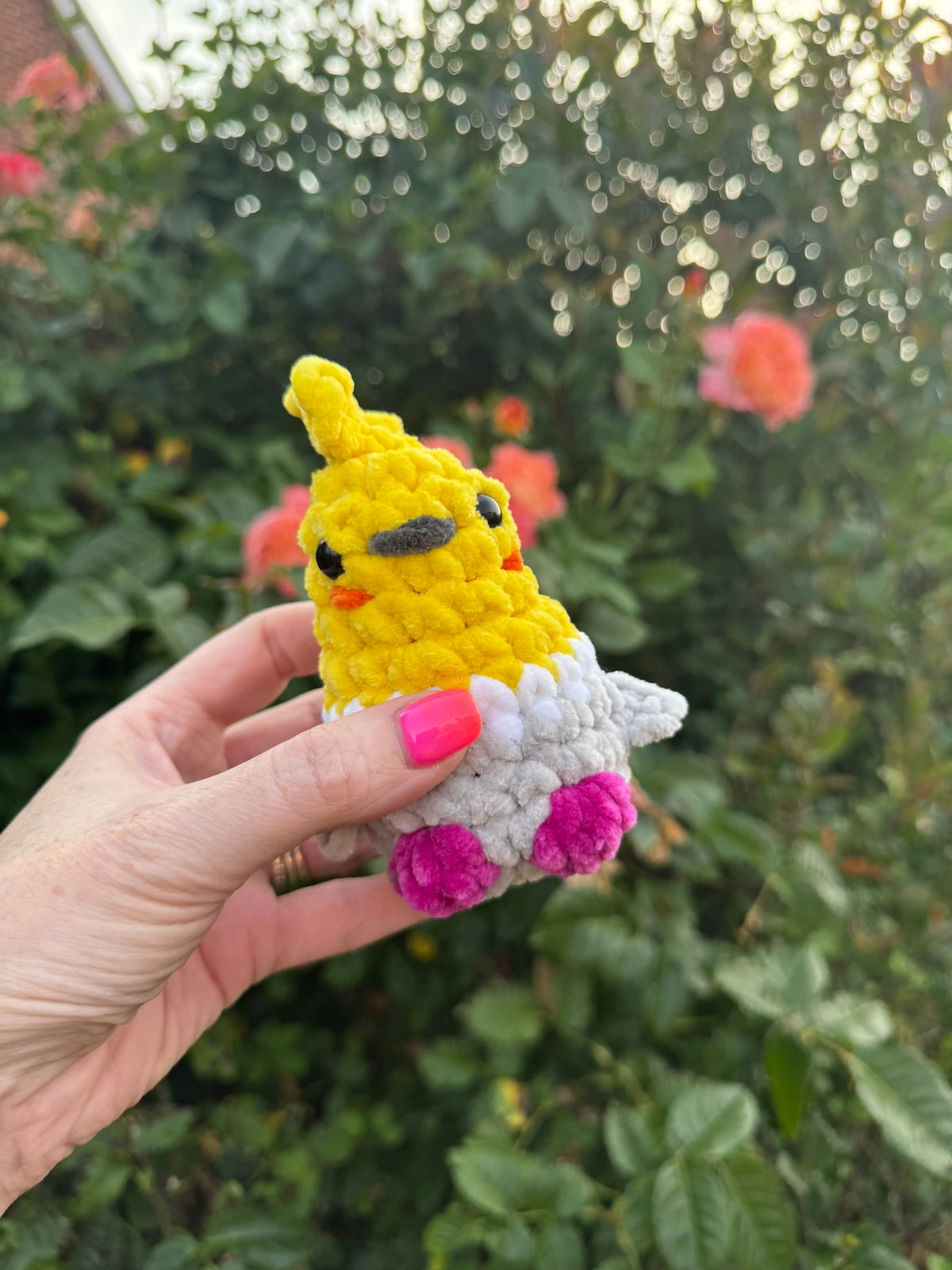 A hand with pink nail polish holds a small crocheted amigurumi chick toy against a blurred garden background of green leaves and pink flowers.
