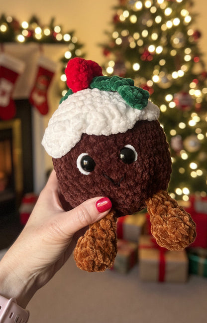 Hand holding a small crocheted Christmas pudding toy with a blurred indoor background