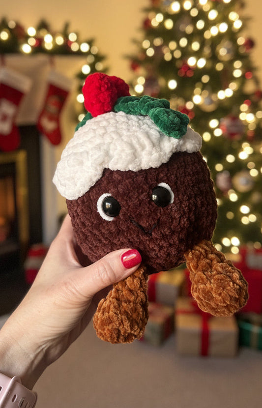 Hand holding a small crocheted Christmas pudding toy with a blurred indoor background