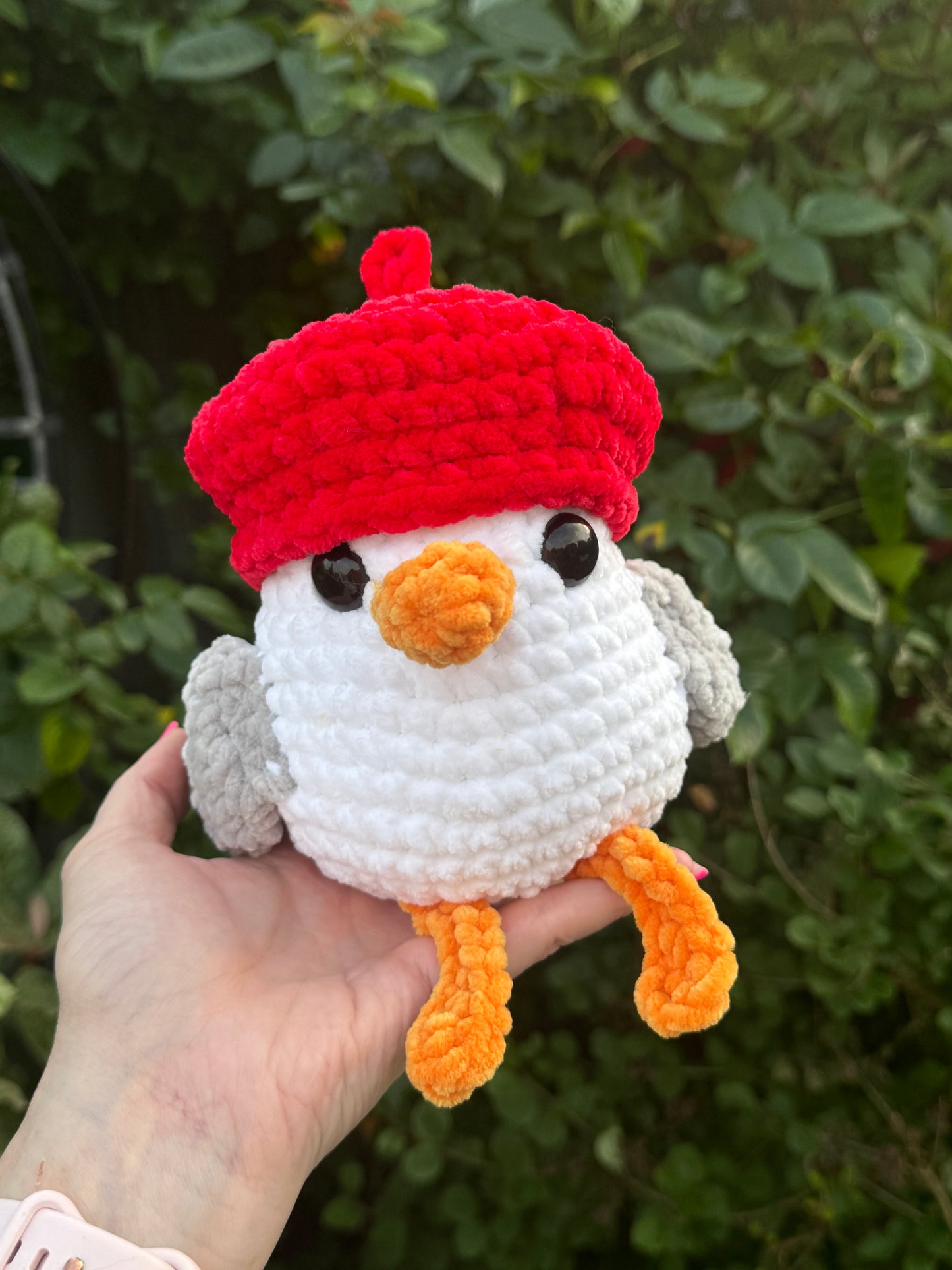 Hand holding a small crochet seagull toy with a red hat against a green leafy background