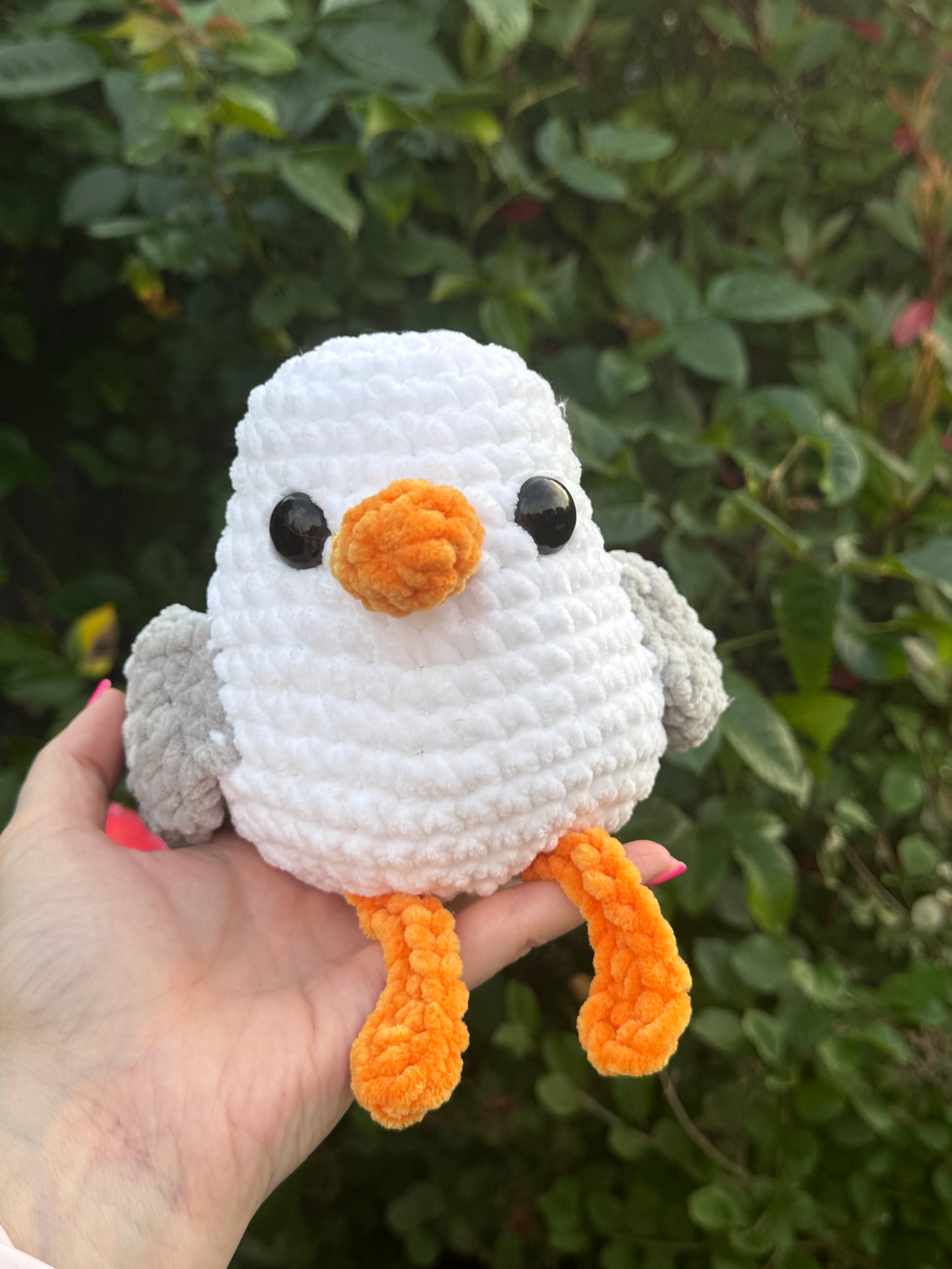 Hand holding a small crochet seagull with a white body, orange beak, and feet against a green leafy background.