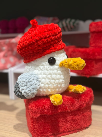 Crocheted bird plush toy with a red hat sitting on a red box.