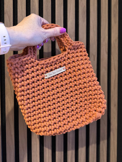 Hand holding a terracotta knitted bag with a brand label against a striped background