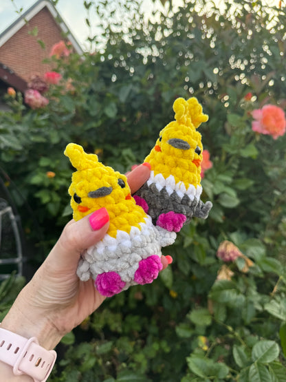 A hand with pink nail polish holds two small crocheted chick toys in front of a blurred garden background with green leaves and pink flowers. The toys are yellow with white, gray, and pink details.