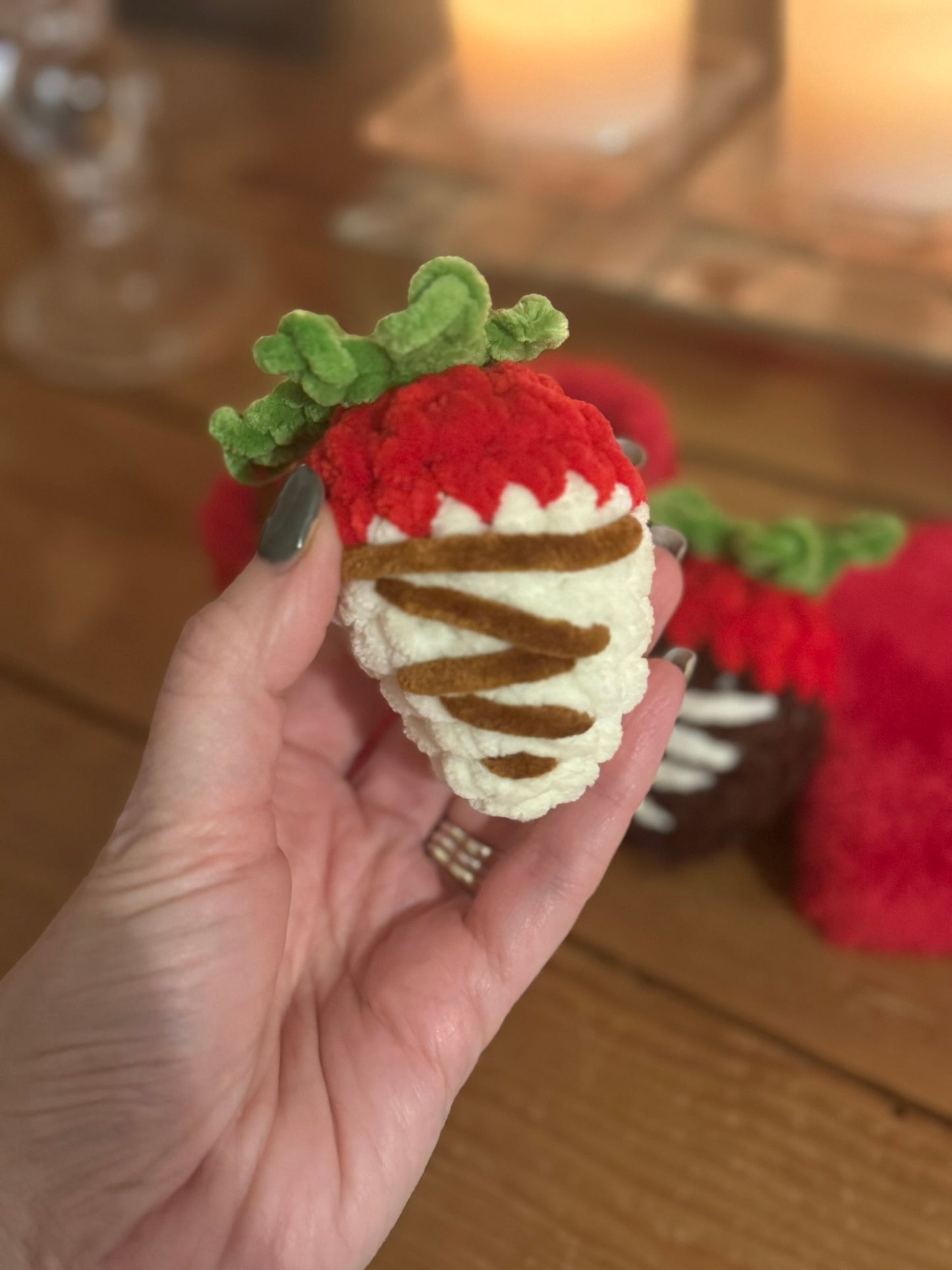 Hand holding a crocheted strawberry with white frosting and green leaves on a wooden surface.